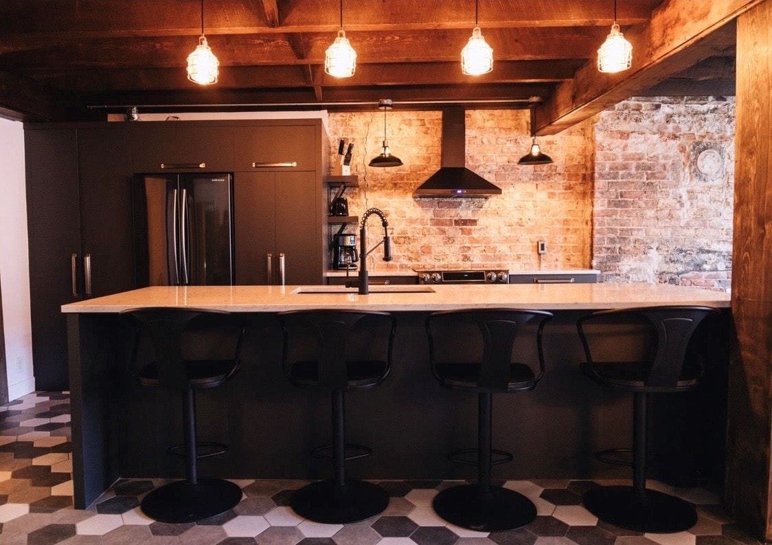 A modern kitchen features a long stone countertop with four black bar stools positioned in front. Exposed brick walls add character, while pendant lights illuminate the space. Sleek cabinetry and stainless steel appliances complete the contemporary design.