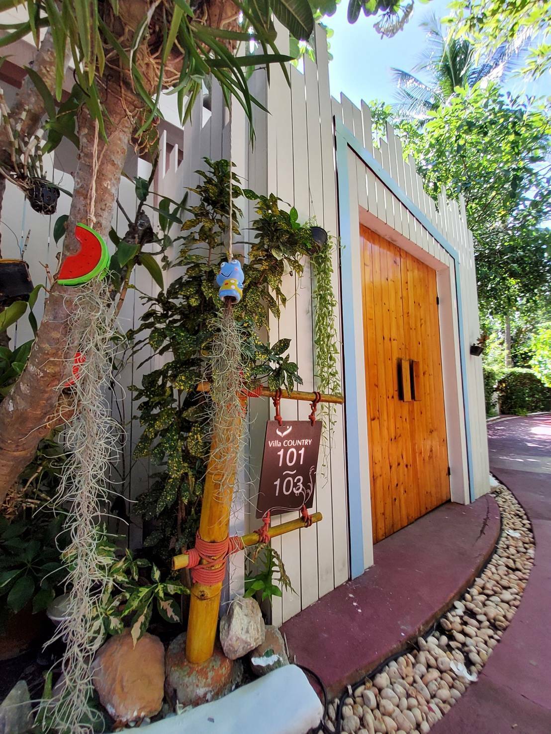 The entrance to the property is framed by lush greenery and tropical plants. A wooden door is visible, accompanied by a sign indicating rooms 101 and 103. Decorative elements, including colorful ornaments and moss, enhance the natural surroundings.