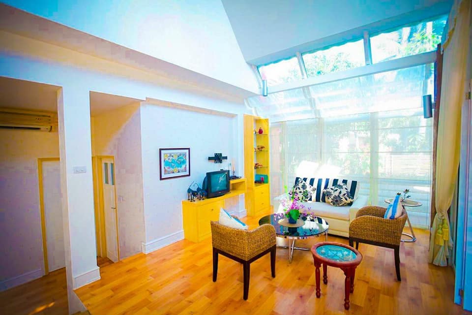 A bright living area features large windows allowing natural light to fill the space. Comfortable seating arrangements include two patterned chairs and a sofa. A small wooden table sits at the center, accompanied by a decorative vase with fresh flowers and a TV against a yellow storage unit.