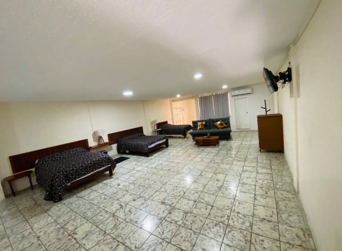 A spacious room features two matrimonial beds with dark patterned bedding, a sofa bed, and a TV mounted on one wall. Natural light flows in through a window, and the tiled floor complements the neutral wall colors, creating a comfortable environment.