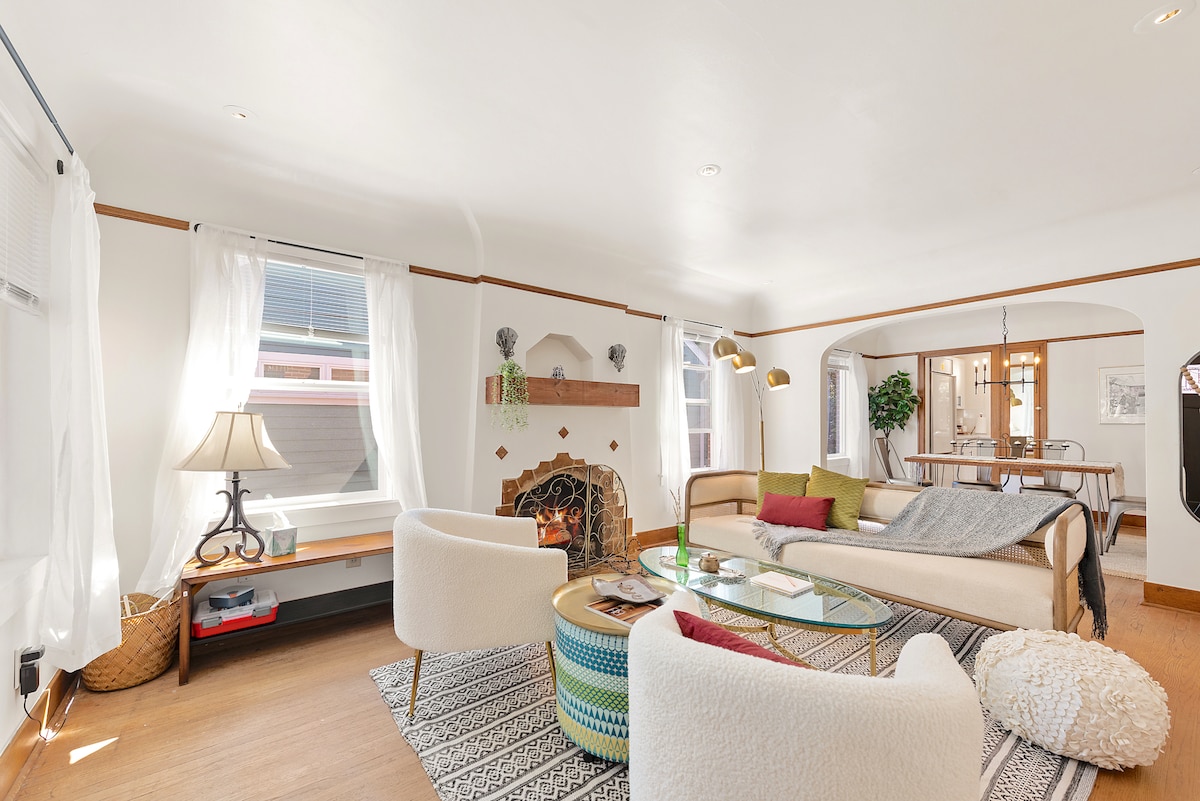 The living room features a cozy seating arrangement with a modern sofa and an elegant glass coffee table. Natural light filters through sheer curtains, and a charming fireplace is framed by decorative elements, creating a warm gathering space. A dining area can be seen in the background.