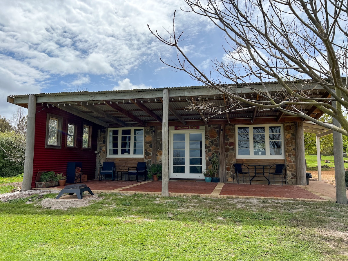 A self-contained cottage is situated on a peaceful landscape, featuring a textured stone facade and a wide porch. Two chairs and a small table are placed outside, offering a welcoming space. Large windows allow natural light into the interior, complementing the serene surroundings.