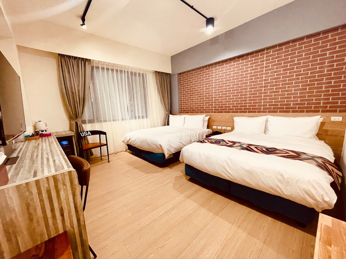 A modern room is shown featuring two spacious beds placed on a light wooden floor. A TV unit can be seen against a textured brick wall, complemented by a writing desk and a chair. Natural light filters through sheer curtains, adding warmth to the environment.