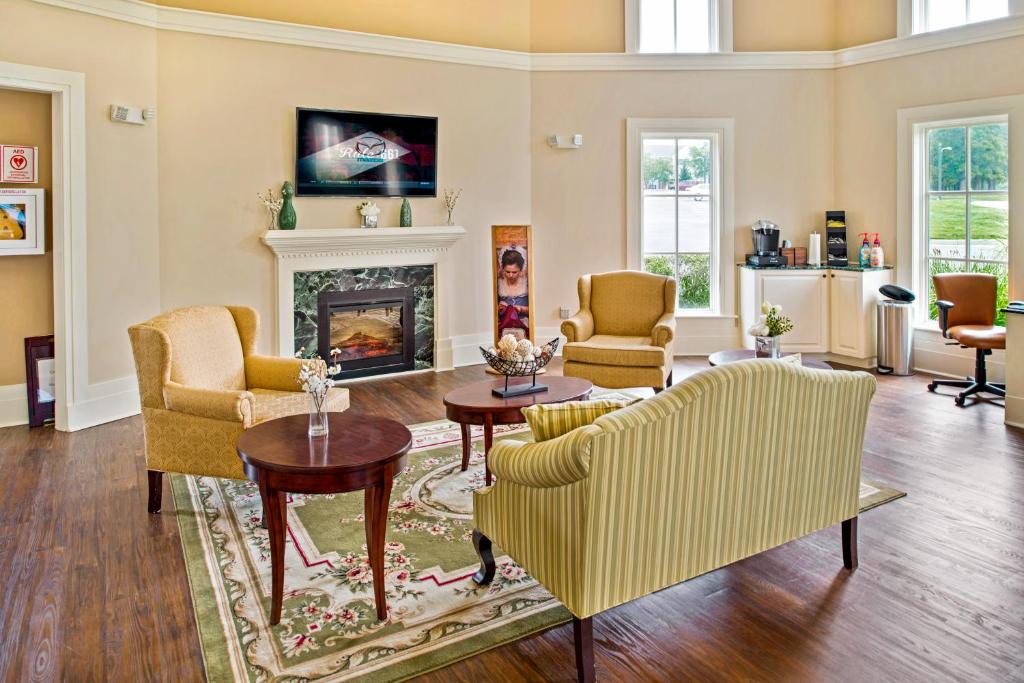 A welcoming common area features a fireplace at the center, with comfortable seating options including two armchairs and a striped sofa. Natural light streams through large windows, and decorative elements such as a floral rug and a television enhance the cozy ambiance.