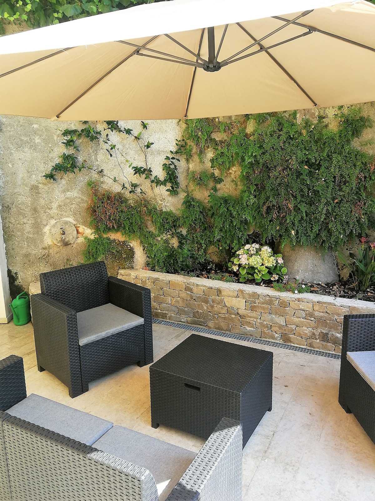 An outdoor relaxation area features four black wicker chairs with soft gray cushions, arranged around a square table. A stone wall is adorned with green foliage and flowering plants, providing a serene backdrop under a large beige umbrella.