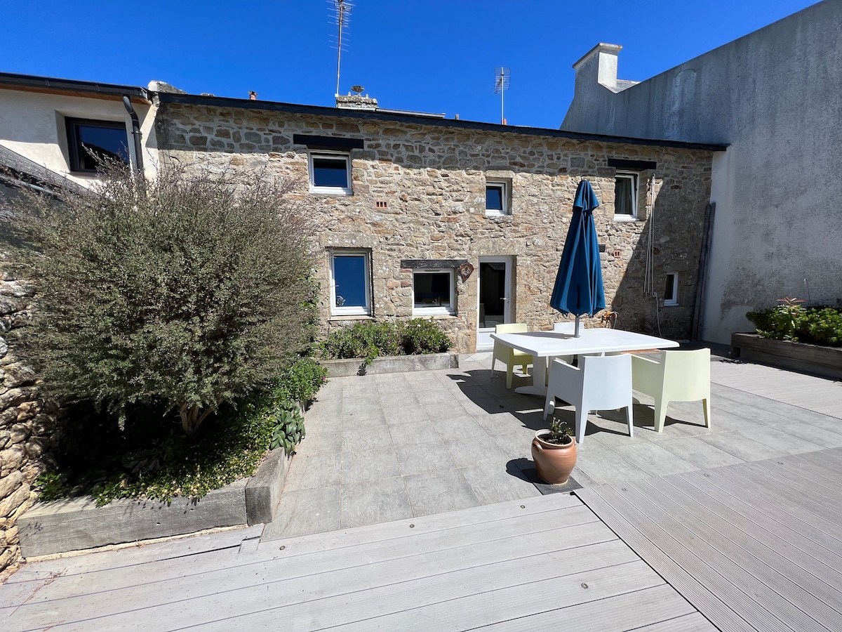 A stone exterior of the house is presented alongside a spacious outdoor terrace. A large white dining table with chairs is set under a blue umbrella. Nearby, lush greenery and potted plants add a natural touch to the inviting space.