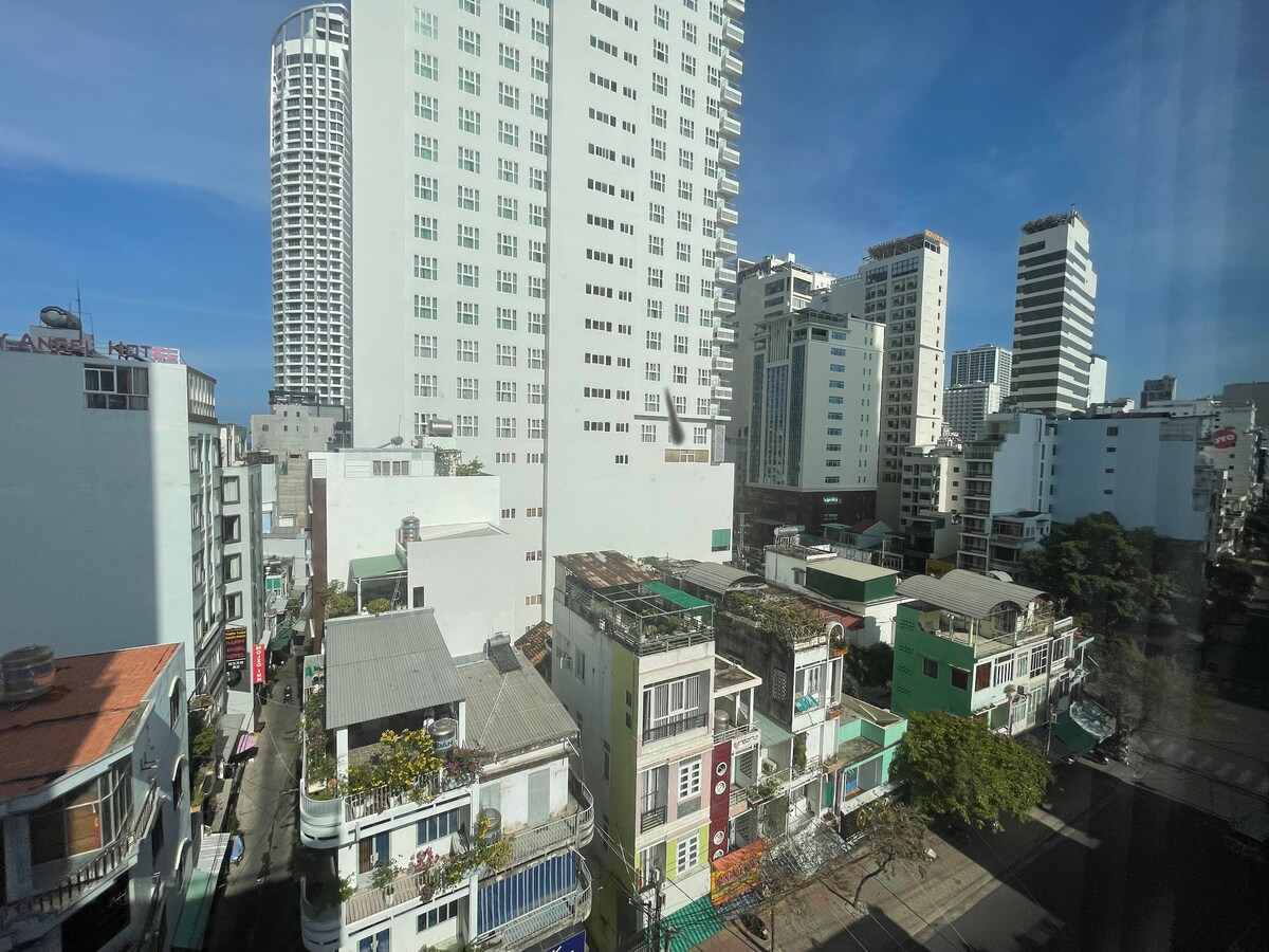 The image captures a view of the surrounding cityscape, featuring a mix of modern high-rise buildings and traditional low-rise structures. Sunlight filters through, highlighting the vibrant colors of the buildings and the bustling urban atmosphere below.