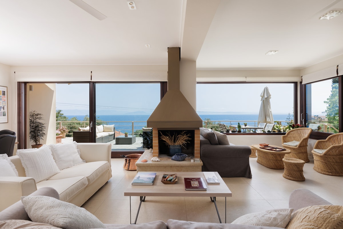 A spacious living room is featured, with large floor-to-ceiling windows providing views of the sea. Comfortable seating arrangements include a mix of light-colored sofas and woven chairs. A central fireplace is present, while a coffee table displays a selection of magazines and books.