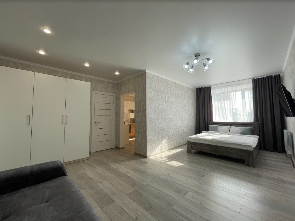 A well-lit interior features a spacious layout with a large window covered by light curtains, allowing natural light to fill the room. A comfortable double bed is positioned beside a modern wardrobe. A soft-toned sofa sits in the corner, enhancing the welcoming ambiance.