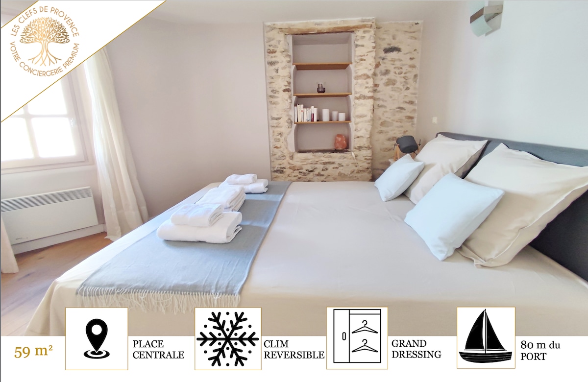 A spacious bedroom features a large bed with clean white linens and an overhead light. A light blue throw is placed at the foot of the bed. The stone wall behind a stylish shelf is complemented by a window allowing natural light to fill the room.