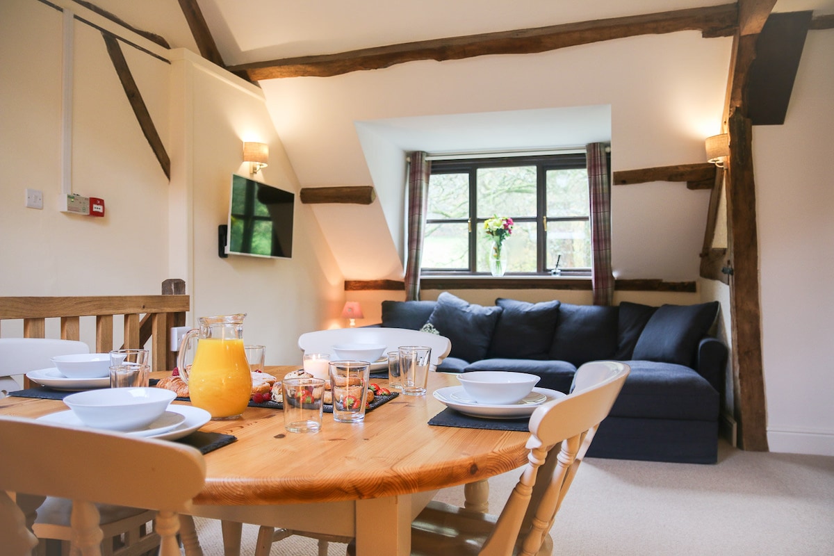 A dining area is presented with a wooden table set for six, featuring plates and glasses. A comfortable sofa is positioned against the wall, with natural light filtering through the window, where a decorative arrangement of flowers is visible.