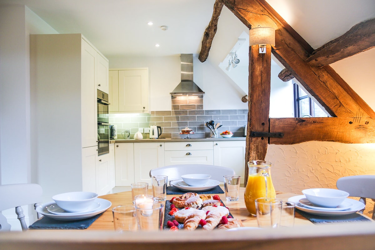 An inviting dining area is highlighted by a wooden table set for breakfast, featuring white dishware and glasses. The modern kitchen is visible in the background, showcasing white cabinetry and a stainless-steel hood. Exposed wooden beams add character to the space.