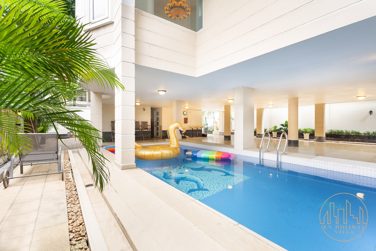 An indoor pool area is featured, showcasing bright blue water and colorful inflatable decorations. Surrounding the pool, a landscaped garden adds greenery, while lounge chairs are positioned nearby for relaxation. Natural light is filtered through large windows, creating an inviting atmosphere.