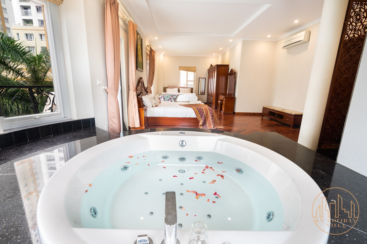 An elegantly designed master bedroom is visible, featuring a spacious indoor setting. A large, circular bathtub is filled with water and adorned with flower petals. Natural light filters through the windows, illuminating the rich wooden flooring and soft furnishings in the background.