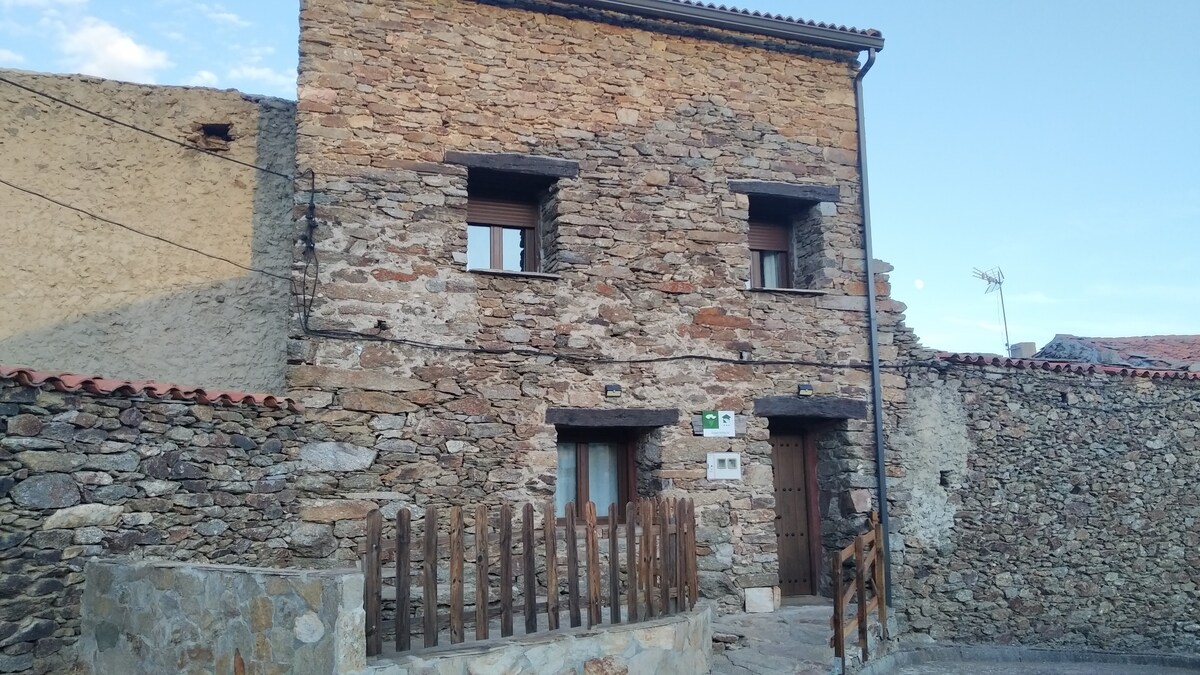 The exterior of a traditional stone house features a rustic façade made of local stone. Two windows are visible on the upper floor, and a wooden door is centrally located on the ground floor. A wooden fence borders a small pathway leading to the entrance.