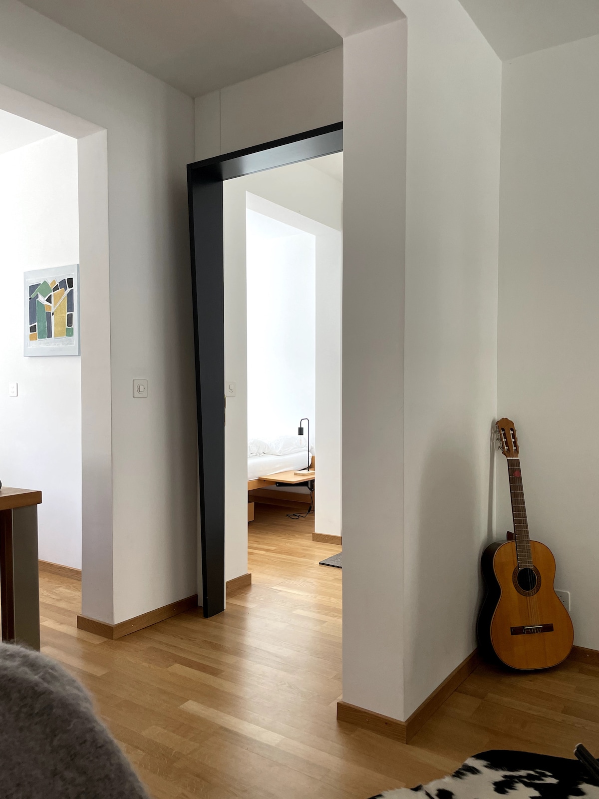 An interior view shows a light-filled passage framed by a sleek black archway, leading into another room. Wooden flooring contributes to a warm aesthetic, while a guitar leans casually against the wall. A glimpse of a bedroom is visible in the background.