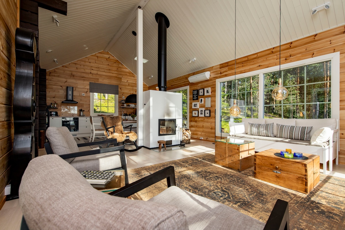 The spacious living area features wooden walls and a high ceiling, with large windows allowing natural light to illuminate the space. A fireplace is situated centrally, surrounded by comfortable seating. Decorative items and artwork add character to the inviting atmosphere.