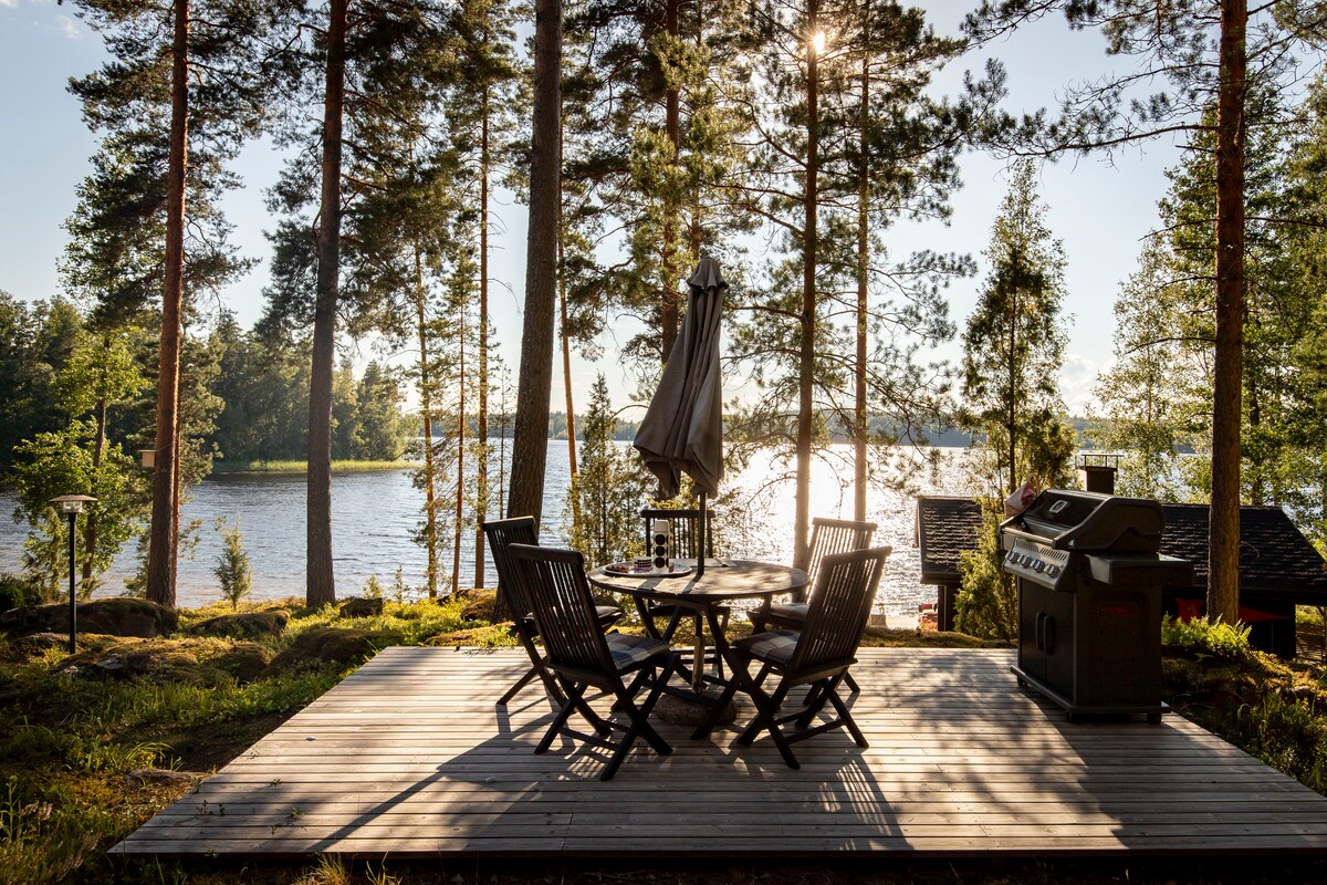 A wooden deck features a round table surrounded by four chairs, with an umbrella positioned at the center. A grill stands nearby, and the view showcases a serene lake framed by tall pine trees and gentle sunlight reflecting off the water.