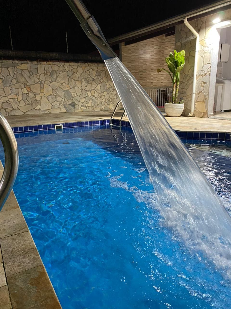 A solar-heated swimming pool is featured, showcasing clear blue water with a cascading fountain creating gentle ripples. The pool is bordered by stone walls and a tiled deck, with an adjacent potted plant visible in the background.
