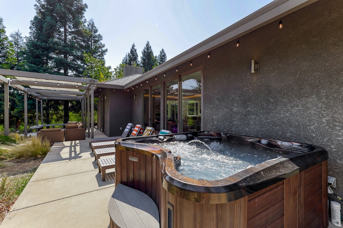 A hot tub is positioned on a concrete patio, surrounded by lounge chairs. Natural light reflects off the water, while landscaping and trees create a serene backdrop. A covered seating area is visible, enhancing the relaxing outdoor space.