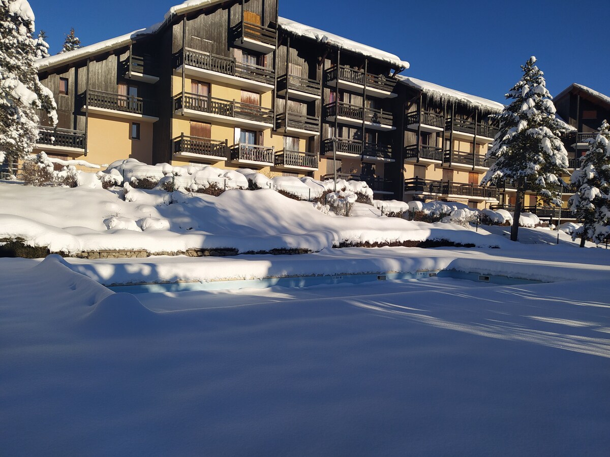 A snow-covered landscape features a pool partially visible, surrounded by a serene blanket of white. The multi-story building stands in the background, with balconies and wooden accents visible among the snow-laden trees.