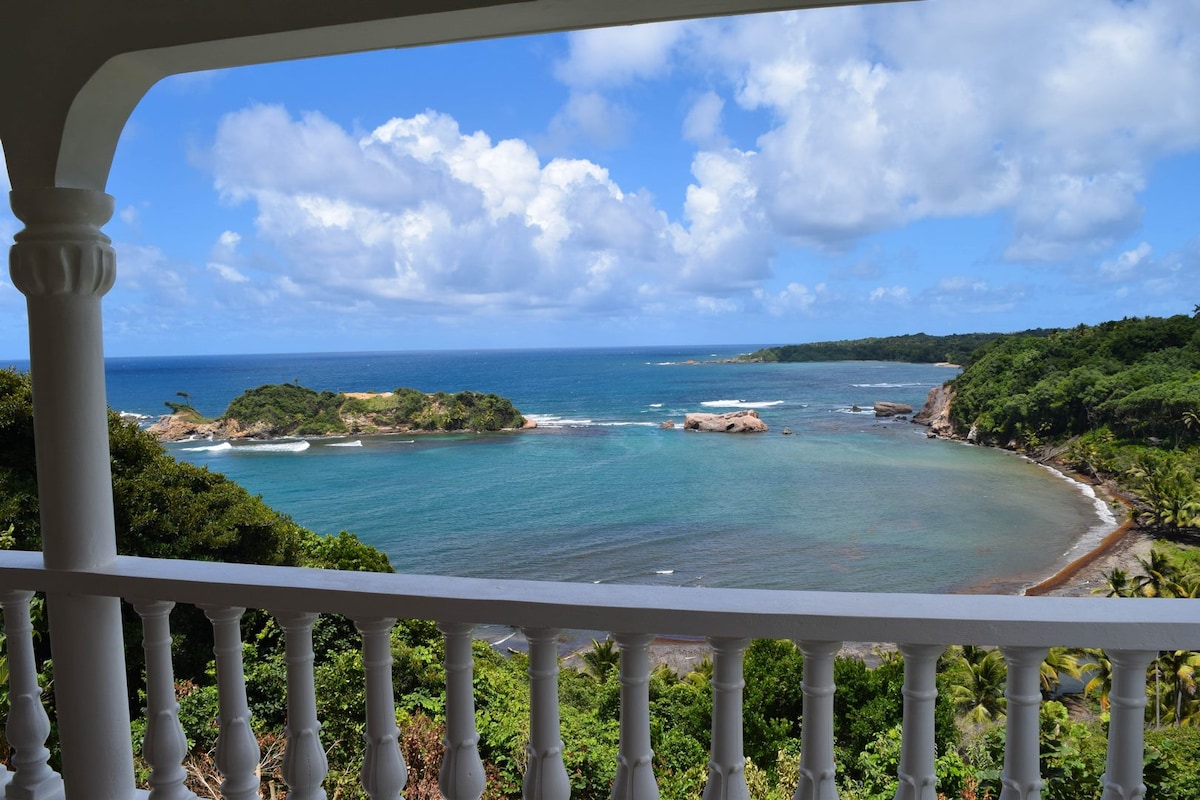 A stunning view of Hodges Bay is captured from the villa's veranda, showcasing calm turquoise waters and lush green coastal hills. The scene is framed by white railings, with scattered clouds in a bright blue sky, enhancing the natural beauty of the surroundings.