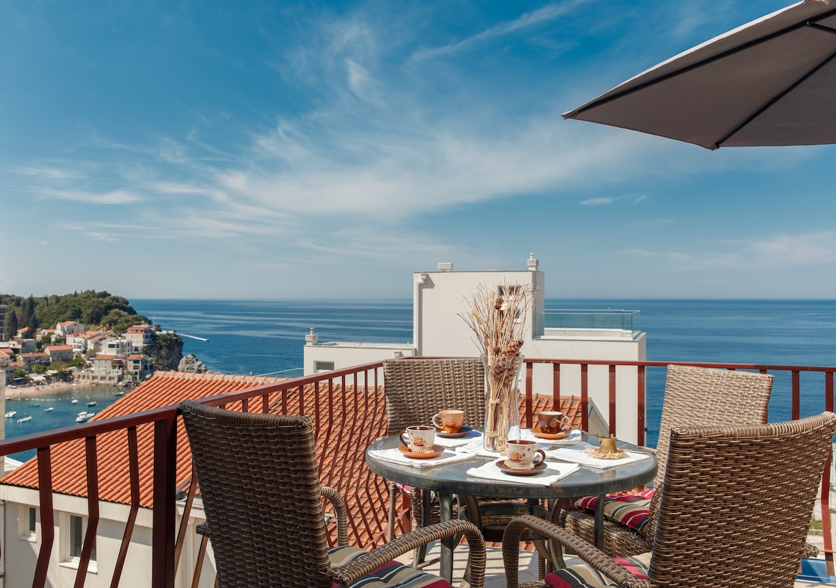 A balcony table is set for breakfast, featuring two cups and plates, surrounded by woven chairs. An umbrella provides shade. Beyond, a panoramic view of the sparkling sea and coastline under a clear blue sky creates a serene setting.