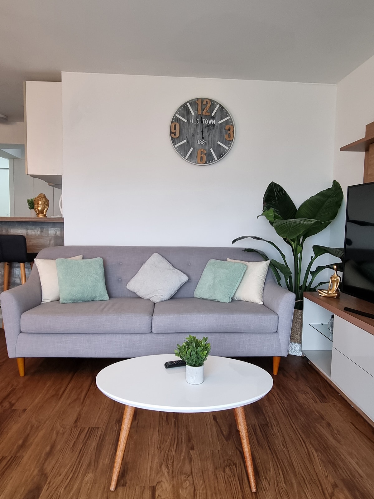 A modern living area is presented with a gray sofa adorned with two decorative pillows. A round coffee table sits in front, accompanied by a small plant. A large wall clock adds a stylish touch above the sofa, with a flat-screen TV positioned nearby.