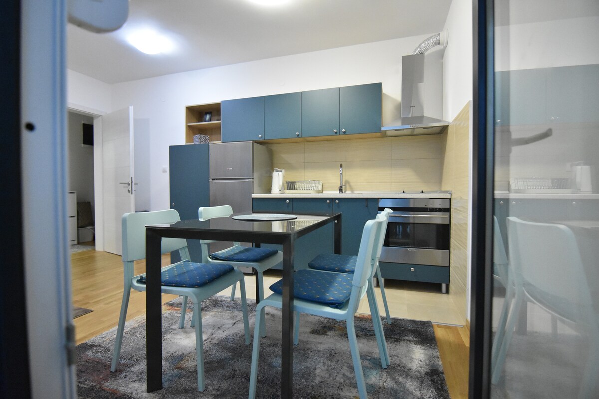 A modern kitchen is displayed, featuring blue cabinets, a sleek stove, and a dining area with a table and four chairs. The space is illuminated by overhead lighting, creating a functional cooking environment.