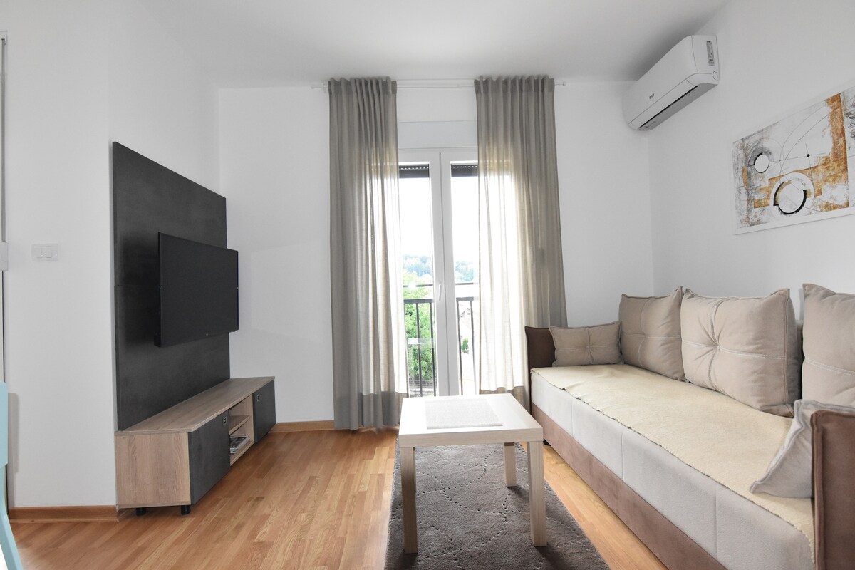 A bright living area is shown, featuring a comfortable sofa with light cushions and a minimalist coffee table. A wall-mounted television is positioned above a wooden media unit. Natural light enters through two large windows adorned with sheer curtains, creating an open and airy feel.