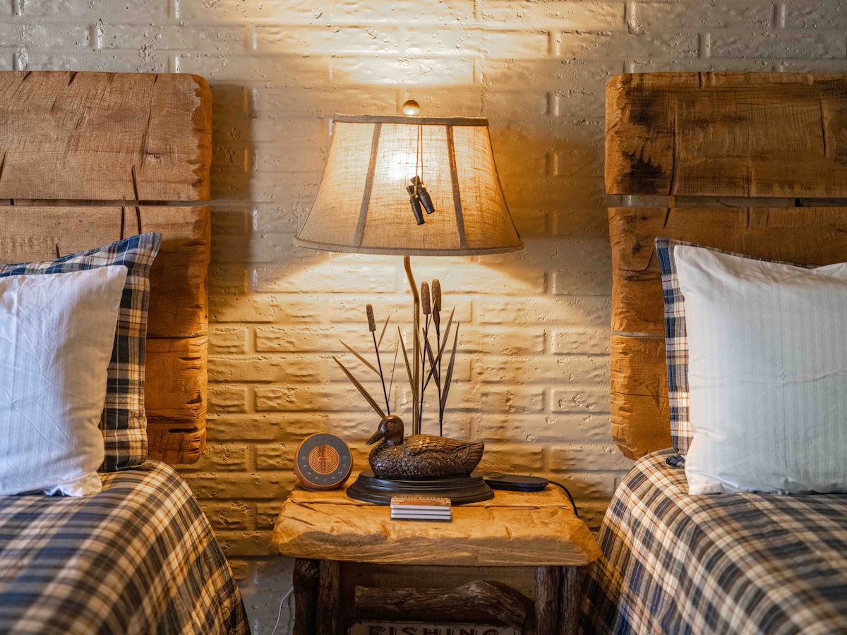 A cozy sleeping area features two plush beds with checkered bedding. Rustic wooden headboards provide a vintage touch. A bedside table hosts a decorative duck and reed accents, alongside a lamp casting warm light against a textured white brick wall.