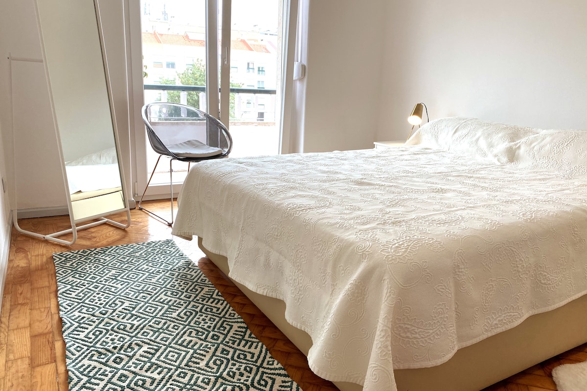 A spacious sleeping area features a large bed with a light-colored coverlet. A cozy chair is positioned nearby, and a full-length mirror stands against the wall. Natural light enters through the balcony doors, illuminating the wooden flooring and geometric rug.