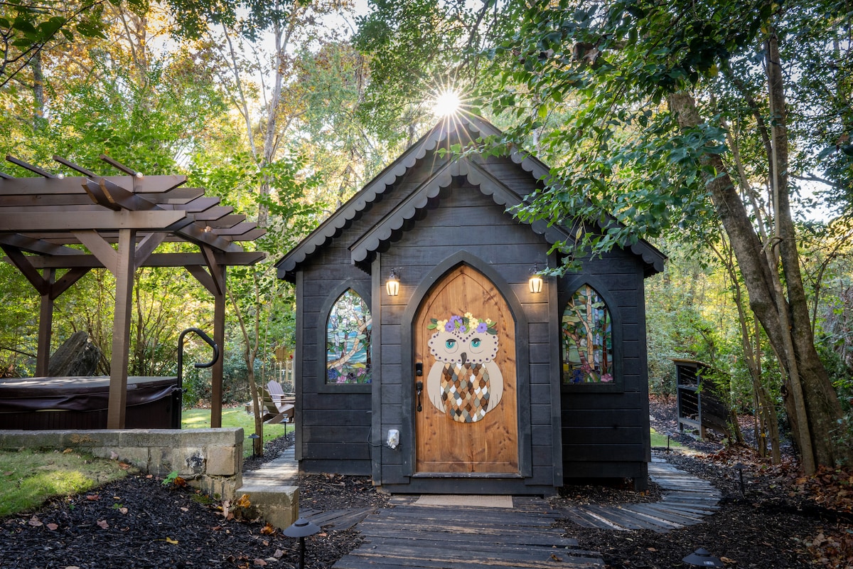 The exterior of a uniquely designed chapel is displayed, featuring an ornate wooden door with owl motifs. Surrounding greenery adds to the secluded ambiance, with sunlight streaming from above. A hot tub and outdoor seating area can be seen beside the entrance, inviting relaxation.