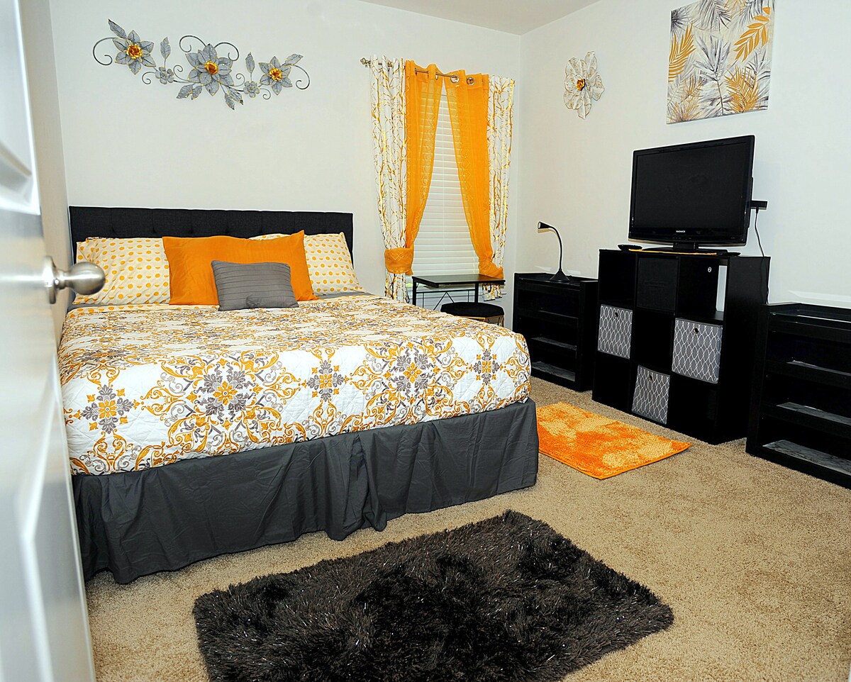 A cozy sleeping area features a spacious bed covered in a patterned duvet with coordinating cushions. Bright yellow and gray accents are present in the curtains and wall decor. A television is positioned atop a chest of drawers, accompanied by a small desk and a plush area rug.