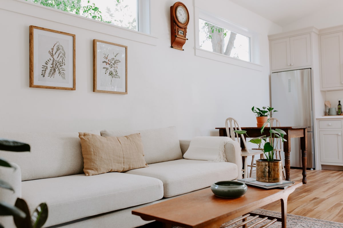 A light-filled living area showcases a comfortable couch adorned with a neutral throw pillow. Two framed botanical prints are mounted on the wall, and a wooden coffee table is set in front. A small dining table and kitchen can be seen in the background.