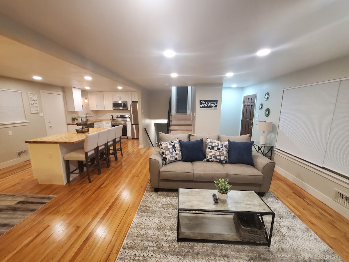 A spacious living area features a comfortable sofa adorned with decorative pillows, paired with a coffee table and a small plant. The warm wood flooring complements the kitchen with bar seating and modern appliances visible in the background. Natural light enters through window treatments.