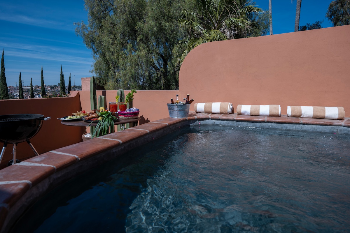 A private terrace is showcased, featuring a jacuzzi surrounded by a charming wall. The space includes plush cushions along the edge, with a nearby table presenting a variety of snacks and beverages. Lush greenery and tall cypress trees are visible in the background.