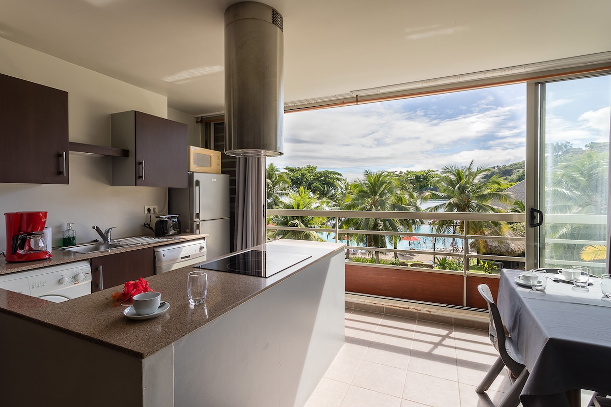 A modern kitchen with dark cabinetry features essential appliances including a stove and refrigerator. A large window provides a view of the palm trees and pool area outside. A dining table set for four stands adjacent to the kitchen, offering a welcoming space for meals.