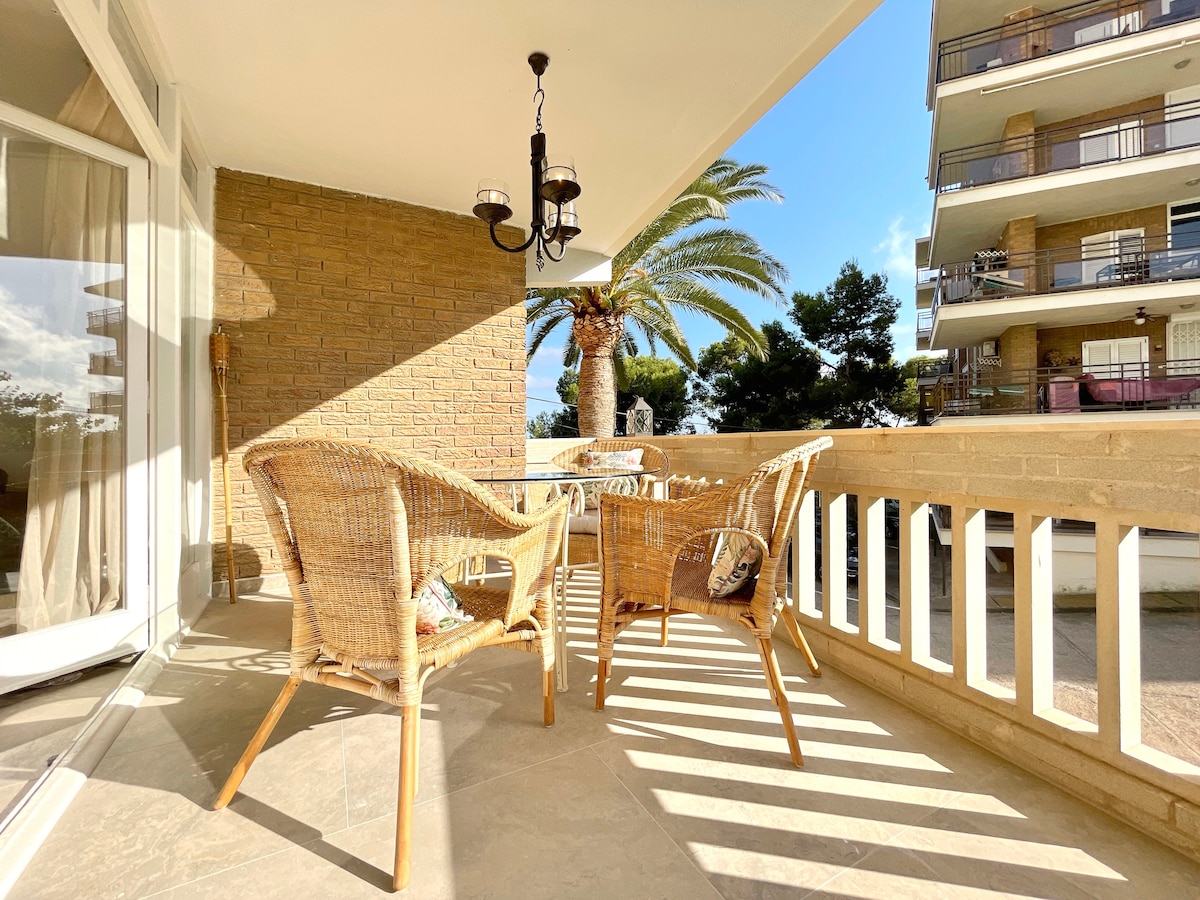 An outdoor terrace features wicker chairs and a matching table, illuminated by natural light. A palm tree is visible in the background, with a glimpse of a multi-story building behind it. Soft shadows create a calming pattern on the stone floor.