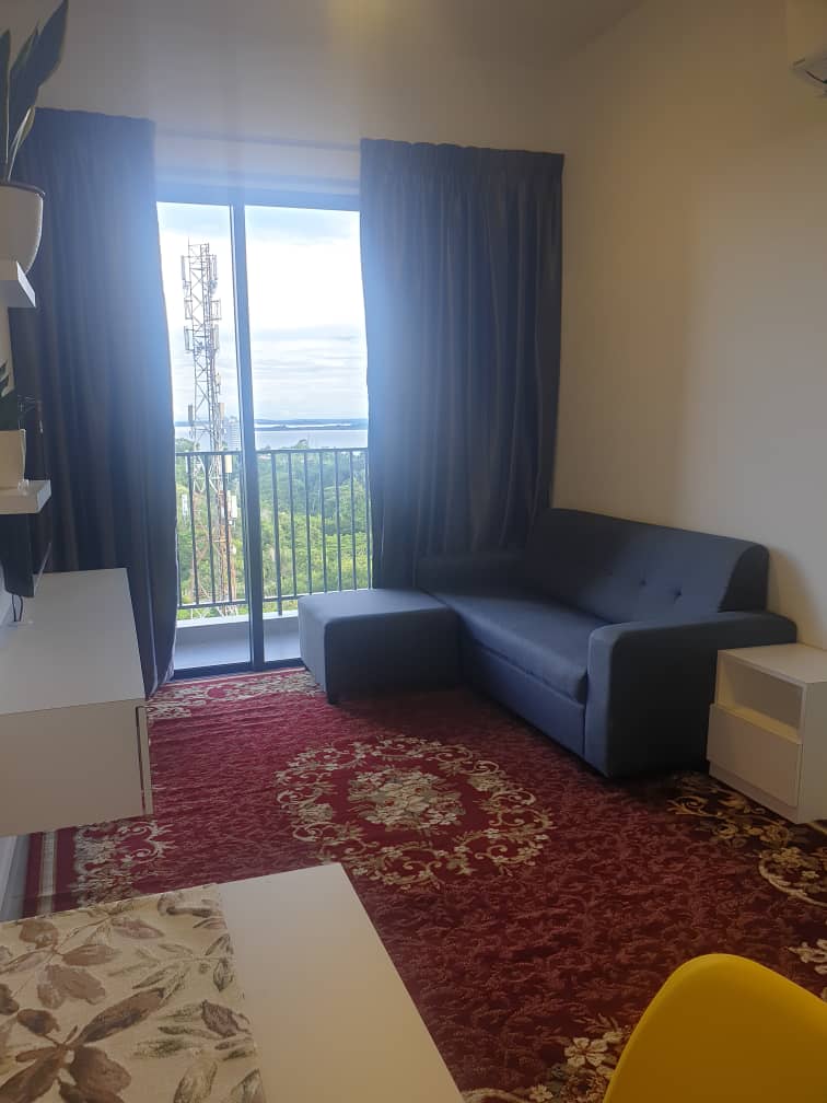A cozy seating area features a gray sofa and a small ottoman on a richly patterned rug. Large windows are draped with dark curtains, offering a view of the surrounding landscape. A white shelf and a desk are visible against the walls.