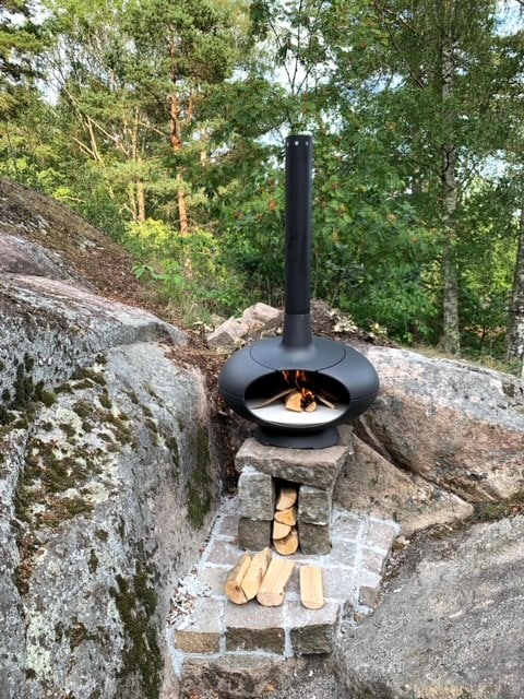 A modern outdoor fireplace is positioned among natural stones and greenery. The sleek black design contrasts with the rough textures of the surrounding rocks. Firewood is neatly stacked next to the fireplace on a stone base, offering an inviting space for warmth and cooking.
