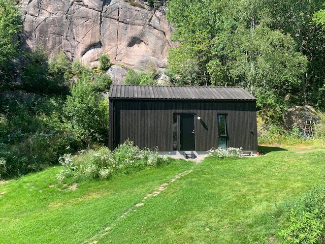 A modern black cottage nestled in a lush green landscape, surrounded by dense foliage and rocky outcrops. The structure features large windows and a double-door entrance, inviting natural light into the space. A stone pathway leads from the grass to the entrance.
