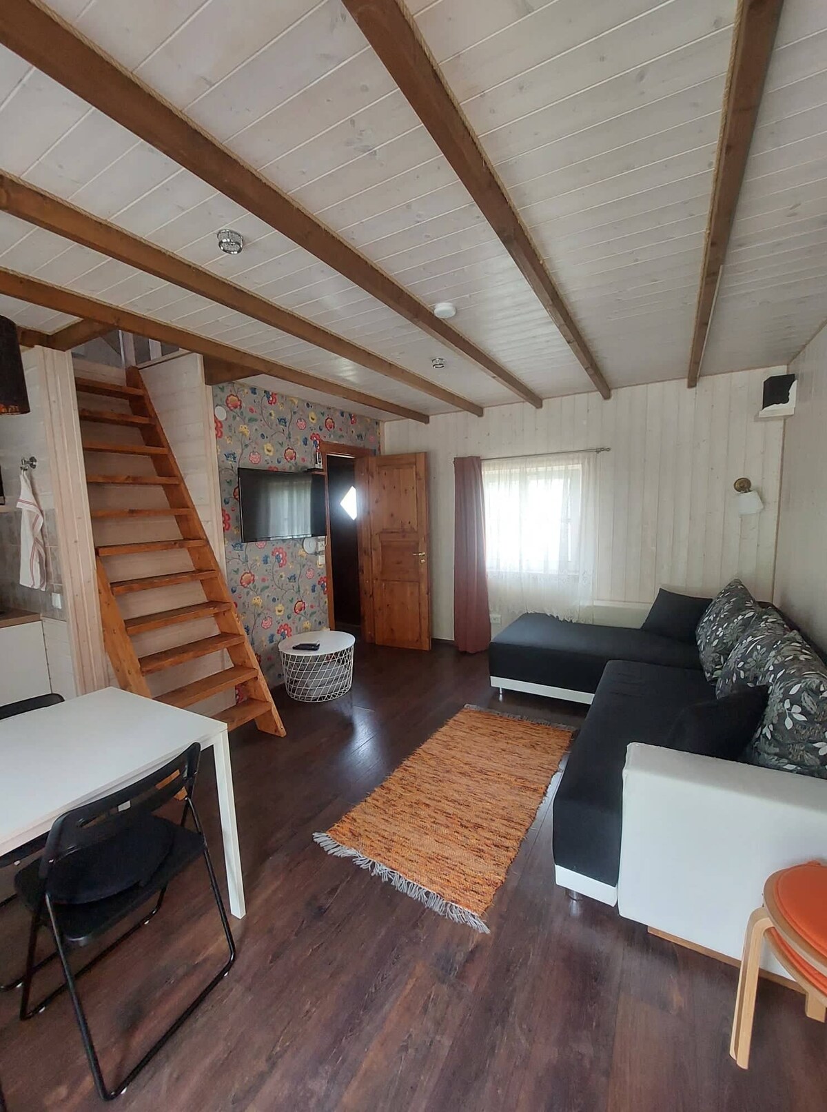An open living area features wooden beams and a staircase leading to an upper level. A comfortable seating arrangement includes a dark sectional couch and a light armchair. A simple dining table with black chairs occupies one corner, while a woven rug adds texture to the floor.