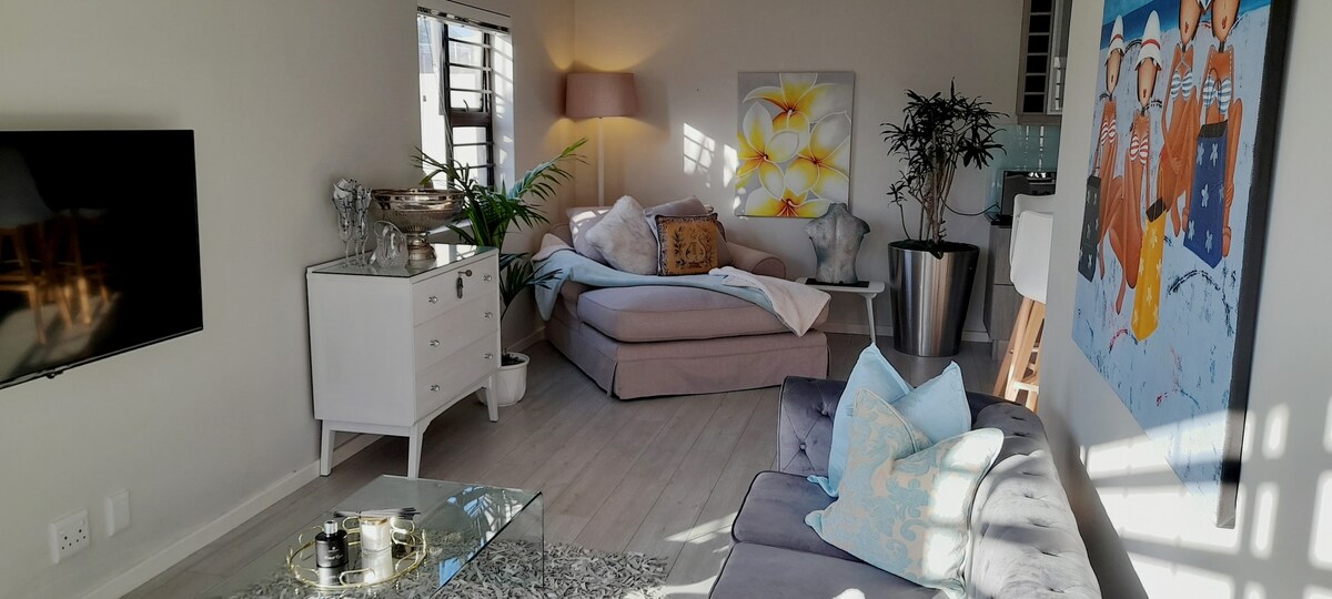 The lounge area features a spacious, light-filled environment with a large sectional couch and a glass coffee table. A TV is mounted on the wall, while a piece of colorful artwork adds interest. Natural light filters through the windows, illuminating a houseplant and decorative elements.