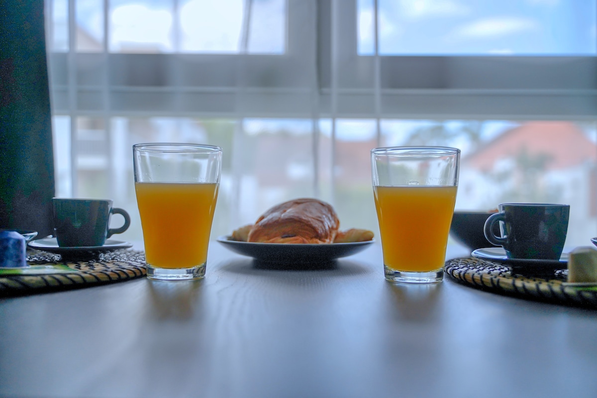 A dining table is set with two glasses of orange juice and two small black coffee cups, alongside a plate of pastries. Natural light fills the room, illuminating the scene through sheer curtains, with a view of the outdoors visible in the background.