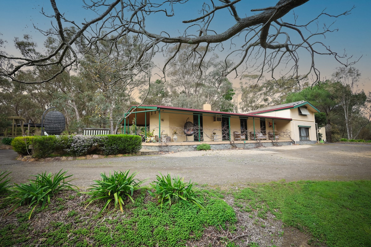 A single-story home is set amidst landscaped gardens, featuring a wide porch with seating areas. Surrounding trees and natural bushland create a serene environment. A gravel driveway leads to the entrance, while greenery adds to the tranquil setting.