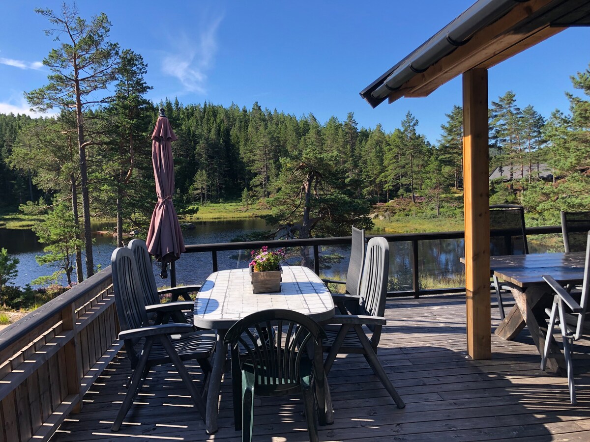 A spacious deck is displayed, featuring a wooden table surrounded by several chairs. An umbrella provides shade, and a potted plant is centered on the table. The serene lake and lush forest create a natural backdrop, enhancing the outdoor setting.