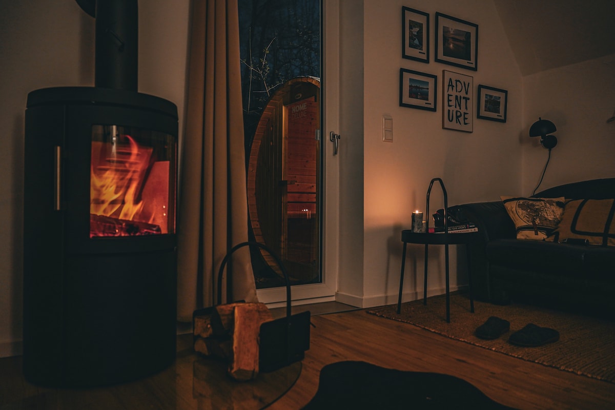 A cozy interior scene showcases a modern wood-burning stove with a visible flame, providing warmth to the room. Soft lighting is present, and a comfortable sofa is adorned with decorative cushions. Nearby, a glimpse of the private sauna is visible through a glass door.