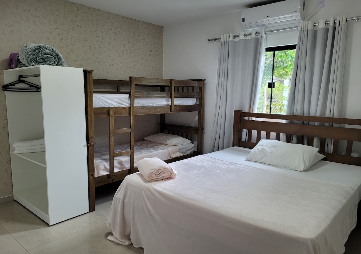 A sleeping area features a spacious bed alongside a wooden bunk bed, accommodating additional guests. Light-colored bedding contrasts with the wooden furniture. A window allows natural light to fill the room, while soft, neutral curtains frame the view. A closet with hangers provides storage options.