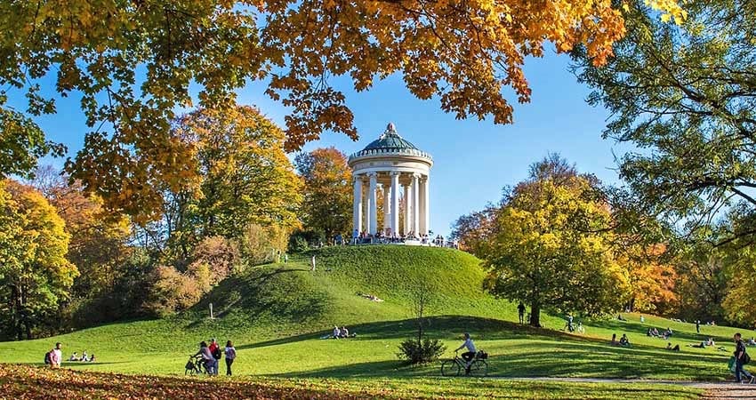 A neoclassical pavilion sits atop a grassy hill, surrounded by colorful autumn foliage. Visitors can be seen enjoying the park, with trees displaying vibrant yellow, orange, and green leaves. The clear blue sky enhances the serene atmosphere of the space.